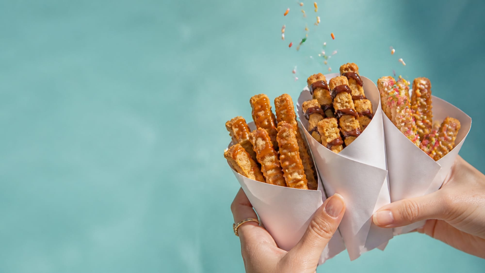 Tres conos de papel con galletas crunch tipo fries bañadas en caramelo y chocolate, con sprinkles cayendo